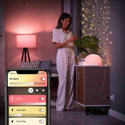 Žena ovláda smart osvetlenie cez mobilnú aplikáciu. Aplikácia zobrazuje: „All lights“, „Shade“, „Table Light“, „Smart Plug“. Kontext: domovské prostredie s nábytkom a svetelnými reťazami.