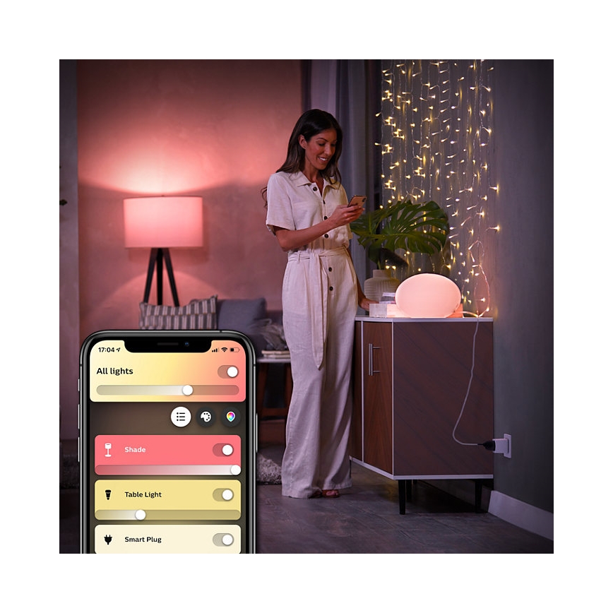 Žena ovláda smart osvetlenie cez mobilnú aplikáciu. Aplikácia zobrazuje: „All lights“, „Shade“, „Table Light“, „Smart Plug“. Kontext: domovské prostredie s nábytkom a svetelnými reťazami.