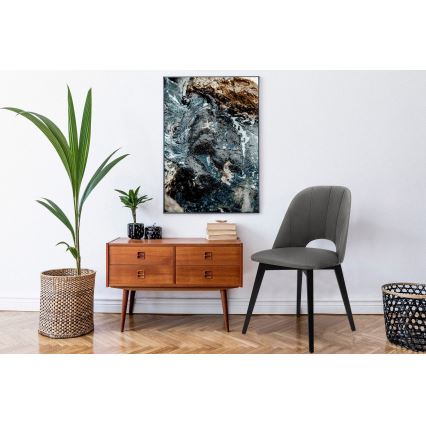 Šedá stolička s tmavými nohami stojí v interiéri. Nachádza sa pred drevenou komodou s obrázkom nad ňou, zobrazujúcim abstraktnú vodnú scenériu tmavomodrých, sivých a hnedých tónov. V okolí sú rastliny a prútený kôš.