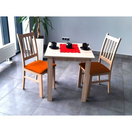 Drevený stôl s dvoma drevenými stoličkami s oranžovými sedákmi stojí na sivej dlažbe. Na stole je červený obrus a dva šálky. V pozadí je rastlina a časť okna.