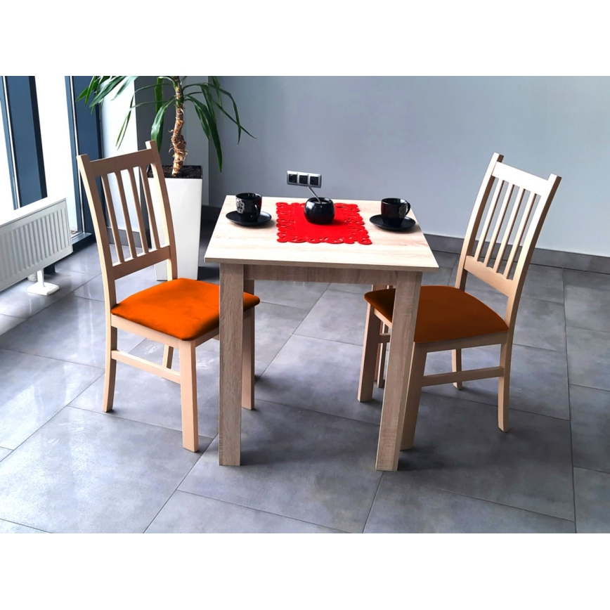 Drevený stôl s dvoma drevenými stoličkami s oranžovými sedákmi stojí na sivej dlažbe. Na stole je červený obrus a dva šálky. V pozadí je rastlina a časť okna.