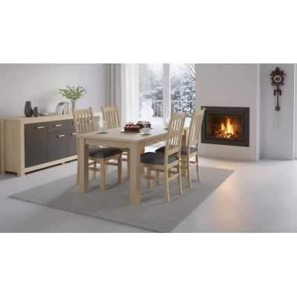 Drevený jedálenský stôl so štyrmi nohami a dvoma stoličkami s tmavosivými sedákmi stojí na tmavosivej dlažbe. Na stole je červený obrus a dve šálky s podšálkami. V pozadí je izbová rastlina a radiátor.