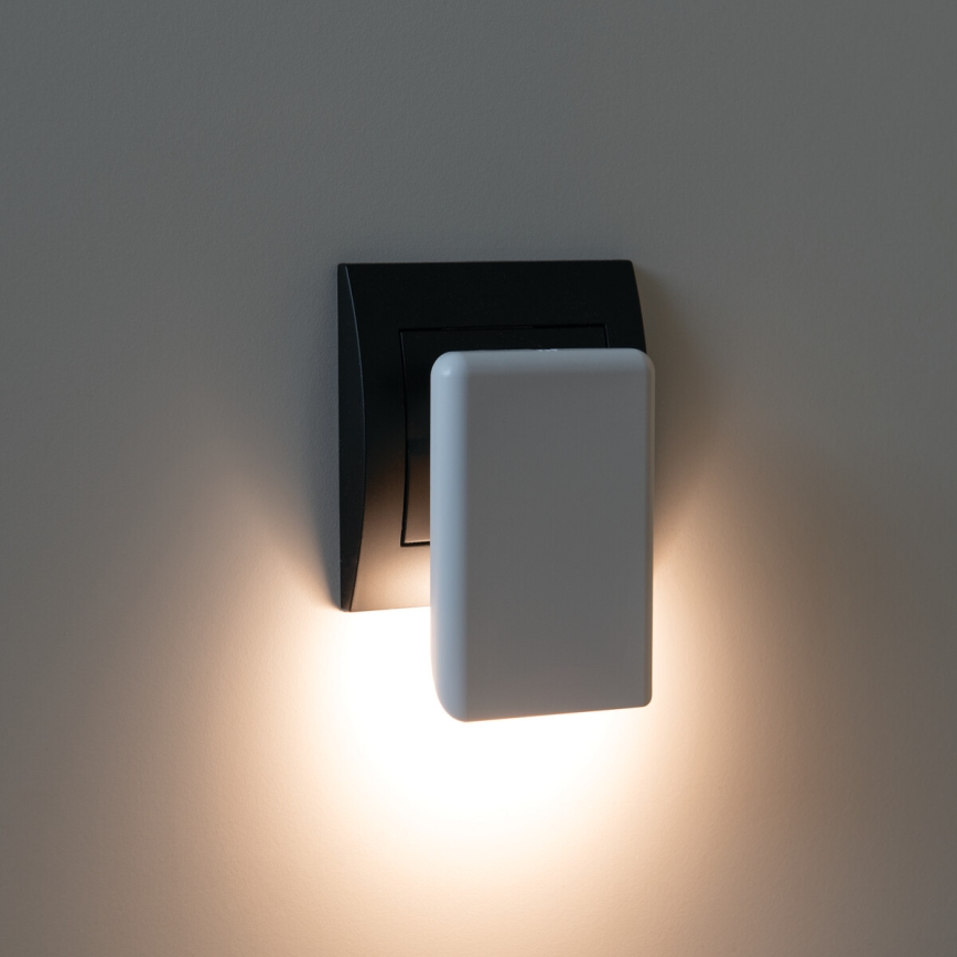 Malá nočná lampa svieti. Je zasunutá do čiernej zástrčky na stene. Svetlo z lampy osvetľuje časť steny.