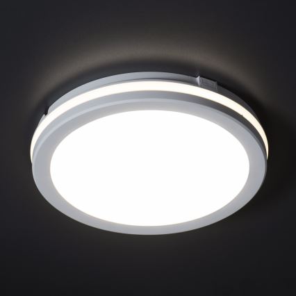 Okrúhle stropné svetlo svieti. Biele svetlo vyžaruje z kruhového difúzora, ktorý je orámovaný dvoma bielymi prstencami. Svetlo je pripevnené k tmavému stropu.