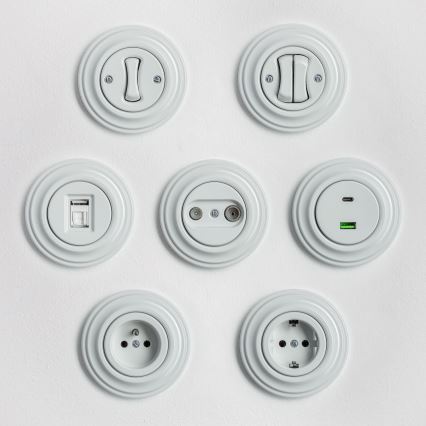 Tri biele vypínače a zásuvka sú umiestnené vedľa seba na bielom pozadí. Dva vypínače sú dvojpolohové a jeden je elektrická zásuvka. Vypínače a zásuvka sú kruhového tvaru s reliéfnym okrajom.