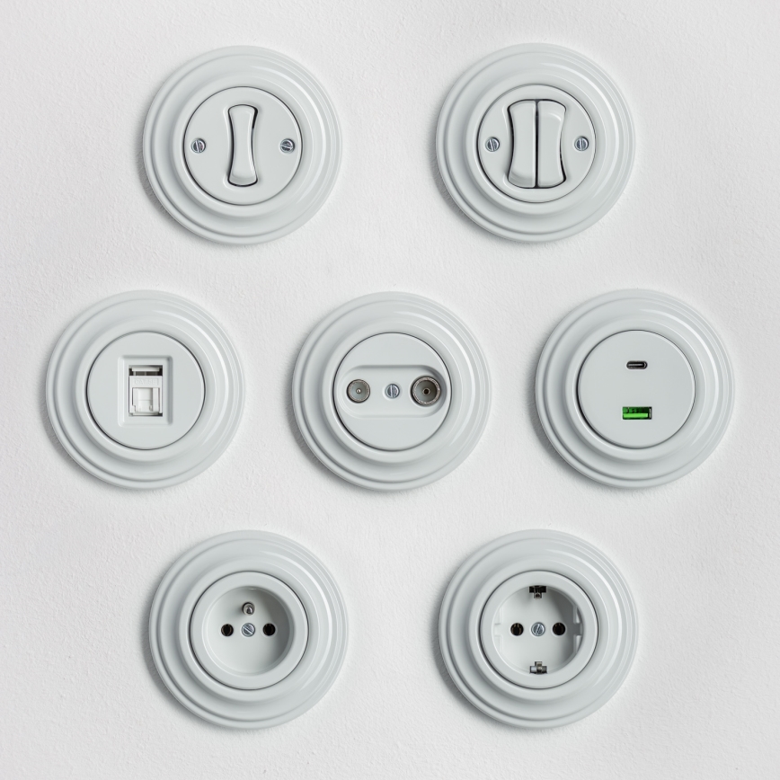 Tri biele vypínače a zásuvka sú umiestnené vedľa seba na bielom pozadí. Dva vypínače sú dvojpolohové a jeden je elektrická zásuvka. Vypínače a zásuvka sú kruhového tvaru s reliéfnym okrajom.