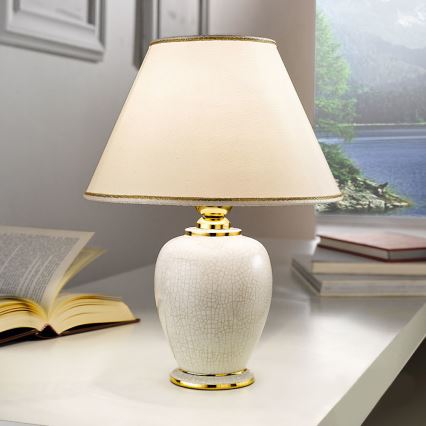 Stolová lampa s krémovou keramickou základňou a bielym tienidlom stojí na bielom stole. Lampa má zlaté detaily. V pozadí sú knihy a okno s výhľadom na jazero.