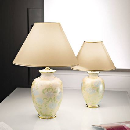 Dve stolové lampy so svetložltými tienidlami a keramickými podstavcami s kvetinovým vzorom stoja na bielom stole. Väčšia lampa je vľavo.