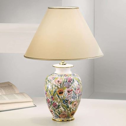 Stolová lampa so svetlo béžovým tienidlom stojí na bielom stole. Jej keramická základňa je zdobená kvetinovým vzorom s prevahou fialiek. Vedľa nej leží kniha.