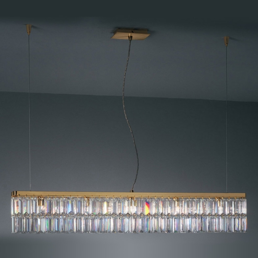 Dlhá zlatá stropná lampa visí zo stropu. Je zdobená mnohými zvislými kryštáľovými prvkami. Lampa je umiestnená pred tmavosivou stenou.