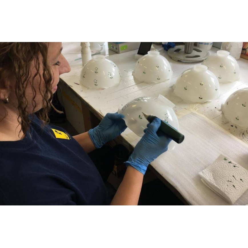 Žena v rukaviciach dekorovala bielu sklenenú misu s tmavozelenou farbou pomocou malého nástroja. Na stole sa nachádzajú ďalšie podobne zdobené misy.