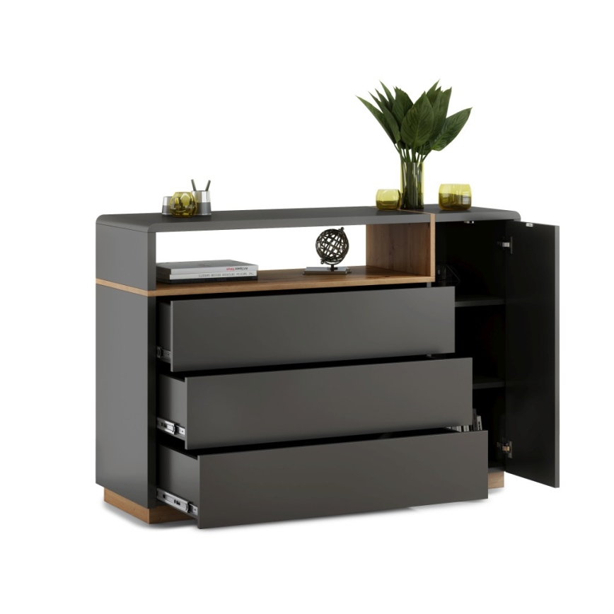Komoda s tromi zásuvkami a otvorenou skrinkou sa nachádza na bielom pozadí. Zásuvky sú tmavosivé, otvorená skrinka obsahuje police. Na hornej poličke komody je umiestnená váza s rastlinami a dekoratívne predmety.
