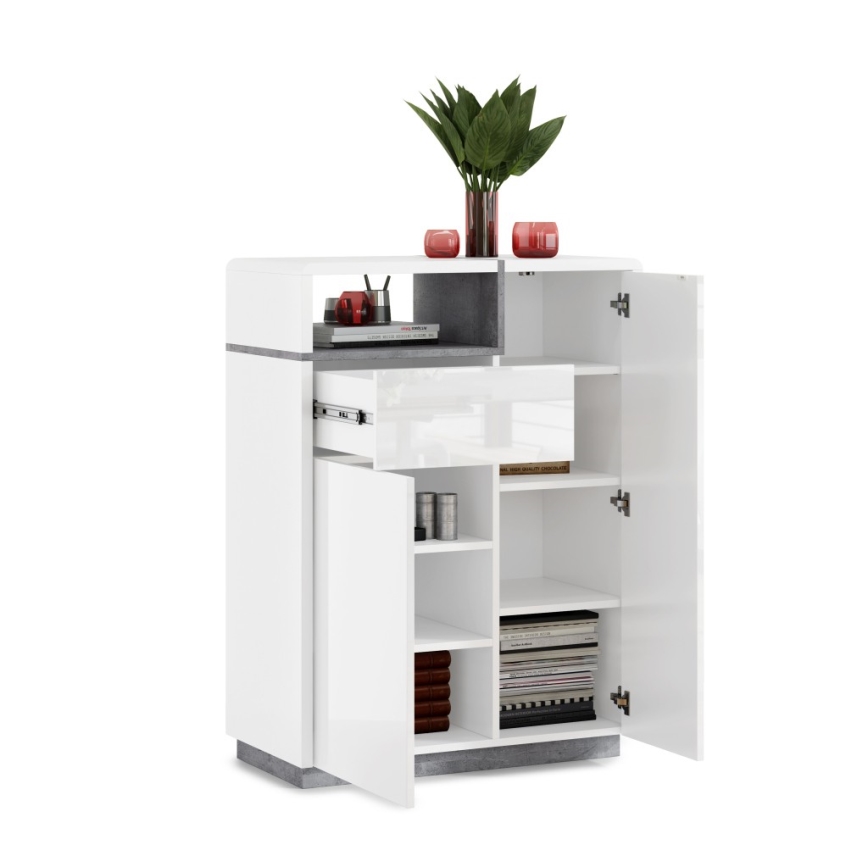 Biely vysoký nábytok s otvorenými dvierkami odhaľuje vnútorné police a zásuvku. Je čiastočne vyplnený predmetmi, ako sú knihy, dekorácie a drobné nádoby. Stojí na bielom pozadí.