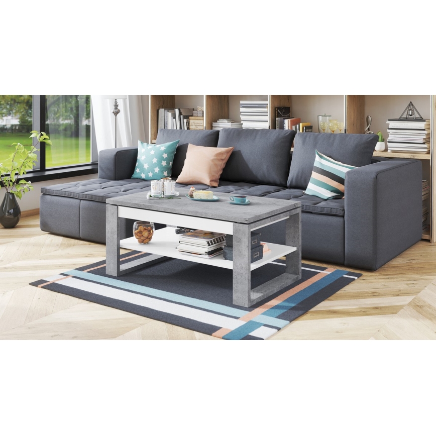 Odkládací stolík s dvoma policami stojí pred sivou rozkladacou sedačkou v obývacej izbe. Stolík je sivý a biely, na policiach sú knihy a dekorácie. Na podlahe je koberec s pruhmi.