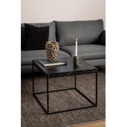 Nízky štvorcový stolík s mramorovou doskou a čiernym kovový rám stojí na tmavosivej koberci pred sivou sedačkou. Na stolíku je kniha a váza s ozdobou a zapálená sviečka v keramickom svietniku.