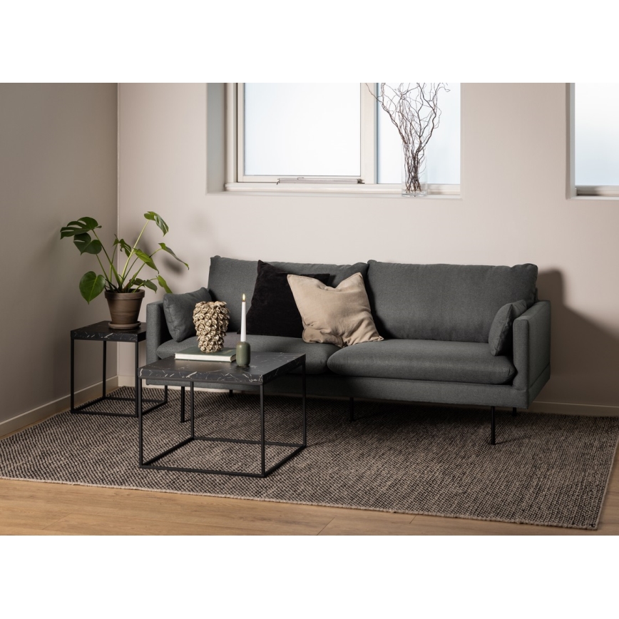 Sivý gauč stojí na tmavošedej koberci v obývacej izbe. Pred ním sa nachádza nízky čierny odkladací stolík z mramoru s kovovou konštrukciou a vedľa menší stolík rovnakého štýlu. Na stolíku je sviečka a dekorácia.