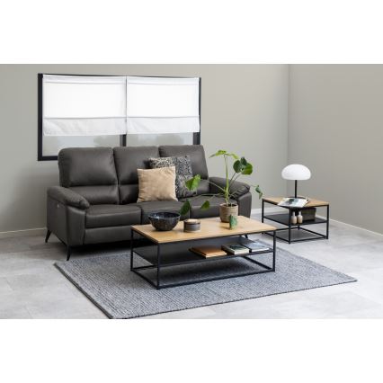 Kovový odkladací stolík s drevenou doskou stojí pred tmavosivou koženou sedačkou. Na stolíku sú dekorácie a knihy. V pozadí je okno s bielymi roletami a svetlosivá stena.