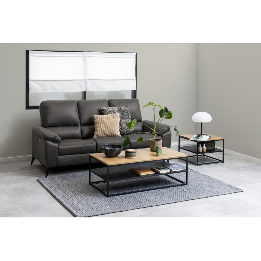 Kovový odkladací stolík s drevenou doskou stojí pred tmavosivou koženou sedačkou. Na stolíku sú dekorácie a knihy. V pozadí je okno s bielymi roletami a svetlosivá stena.