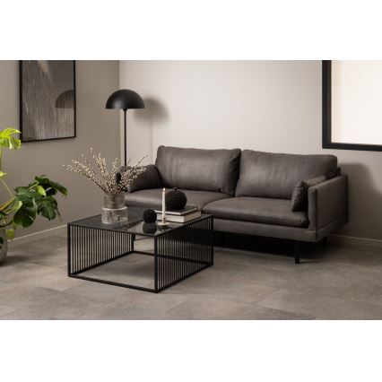 Čierny odkladací stolík so sklenenou doskou a kovovou konštrukciou stojí pred sivou pohovkou. Na stolíku sú dekorácie: váza s konárikmi, sviečka a gule. Pohovka je v obývacej izbe s neutrálnou stenou a podlahovou dlažbou.