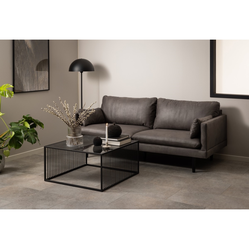 Čierny odkladací stolík so sklenenou doskou a kovovou konštrukciou stojí pred sivou pohovkou. Na stolíku sú dekorácie: váza s konárikmi, sviečka a gule. Pohovka je v obývacej izbe s neutrálnou stenou a podlahovou dlažbou.