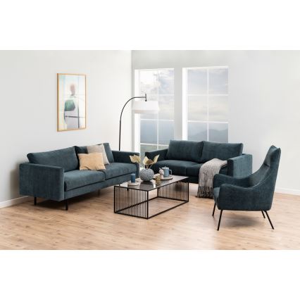 Tmavomodrá rozkladacia sedačka s vankúšmi stojí v obývacej izbe s drevenou podlahou. Popri nej je menšia sedačka a kreslo v rovnakej farbe. V strede je konferenčný stolík s dekoráciami. Izba má veľké okná s výhľadom na krajinu a podlahovú lampu.