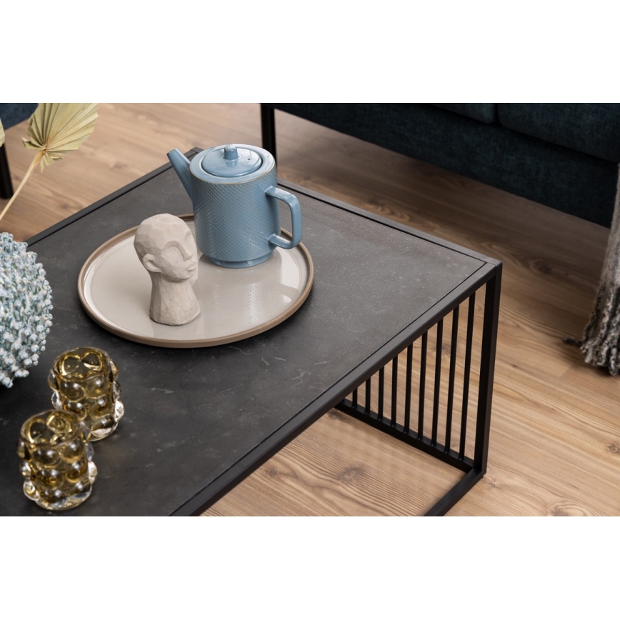 Odporúčam nasledovný opis:Odporúčam nasledovný opis: Nízky obdĺžnikový konferenčný stolík s tmavosivou doskou a čiernym kovovým rámom stojí na svetlohnedej podlahe. Na stolíku sa nachádza svetlohnedá podnos, na ktorom je umiestnená modrá čajová kanvička a malá soška hlavy. Vedľa podnosu sú dva sklenené svietniky.