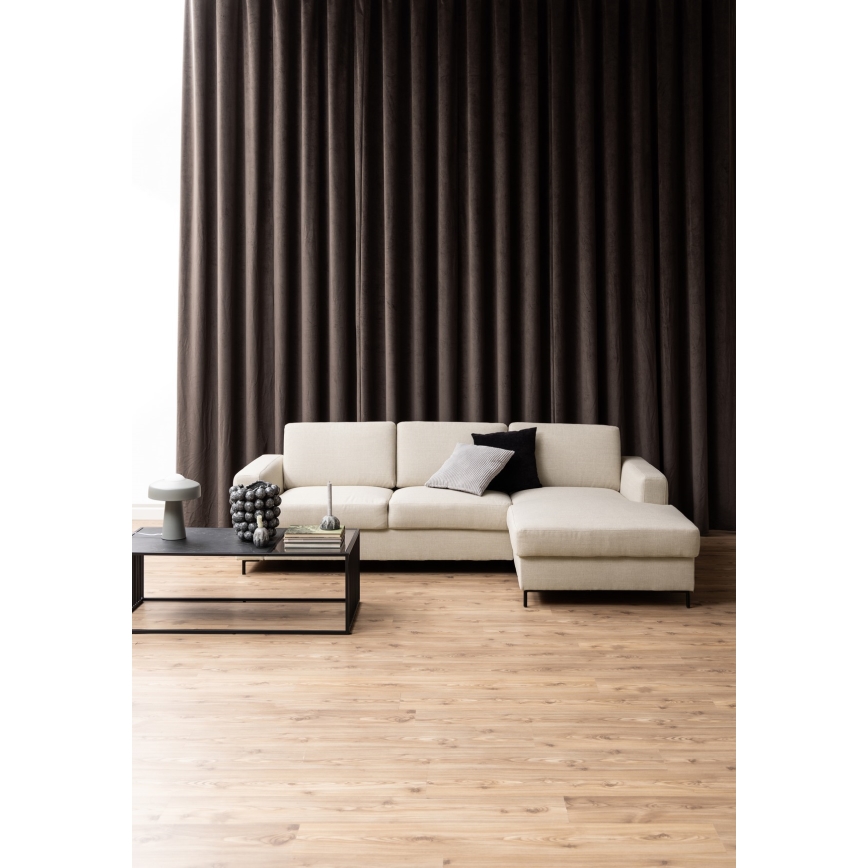 Rohová sedačka svetlo béžovej farby stojí v obývacej izbe pred tmavohnedými závesmi. Na nízkom čiernom konferenčnom stolíku sú dekoratívne predmety. Podlaha je svetlá drevená.