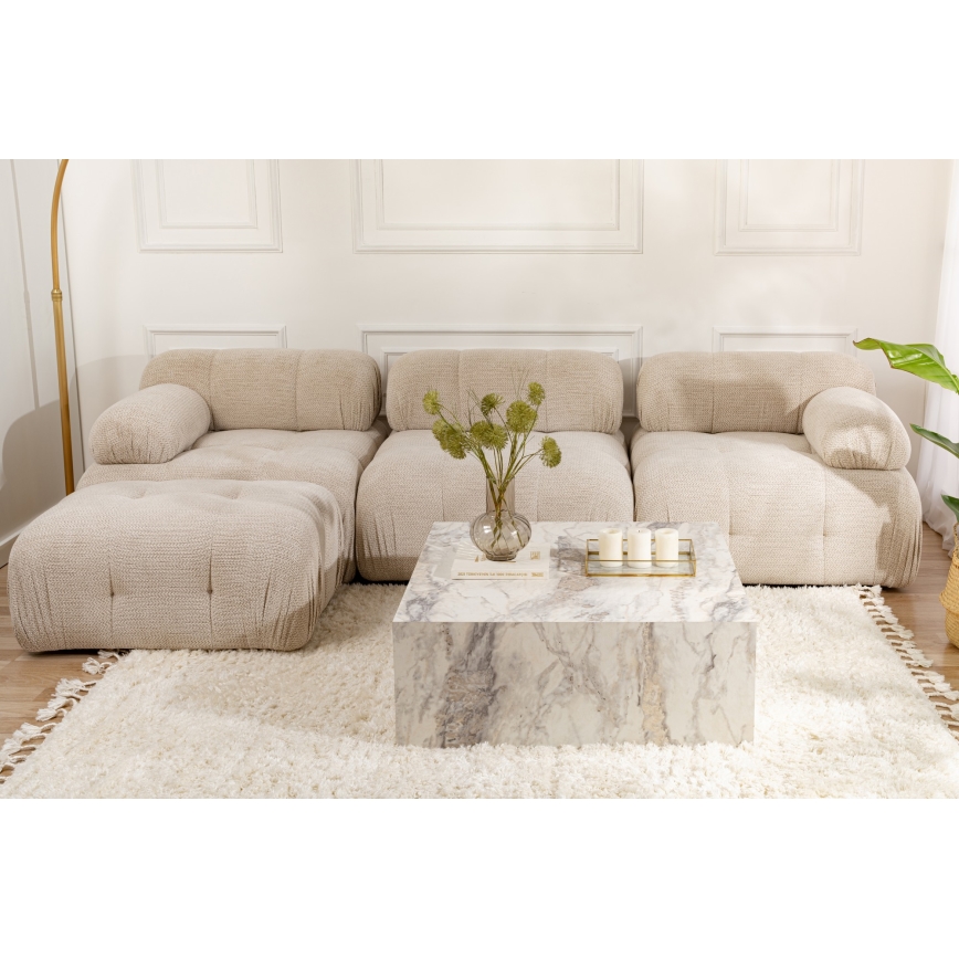 Rohová sedačka svetlo béžovej farby stojí na svetlom koberci. Na nízkom mramorovom konferenčnom stolíku je váza s kvetmi a sviečky. Sedačka je členitá, s výraznými vypchými vankúšmi.