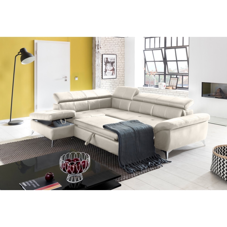 Rohová sedačka svetlo béžovej farby je rozložená v moderne zariadenej obývacej izbe. Sedačka má úložný priestor a je prekrytá tmavosivou dekou. Izba obsahuje aj konferenčný stolík a podlahovú lampu.