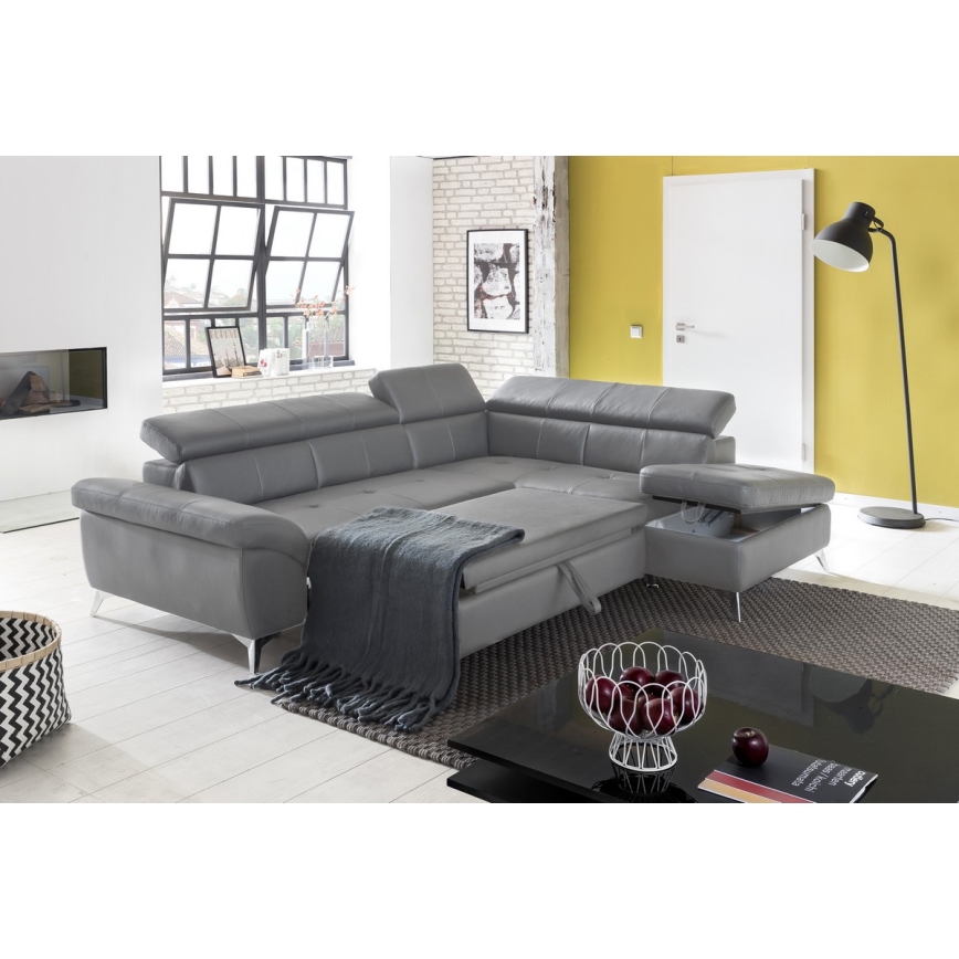 Sivá rohová sedačka s úložným priestorom stojí v moderne zariadenej obývacej izbe. Na sedačke je prehodená sivá deka. V miestnosti je vidieť podlahu, stôl a lampu.