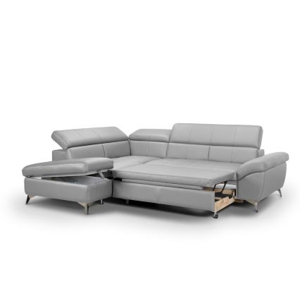 Rohová sedačka svetlosivej kože je rozložená, odhalený je úložný priestor. Súčasťou je aj samostatný taburet s úložným priestorom. Sedačka stojí na svetlom pozadí.