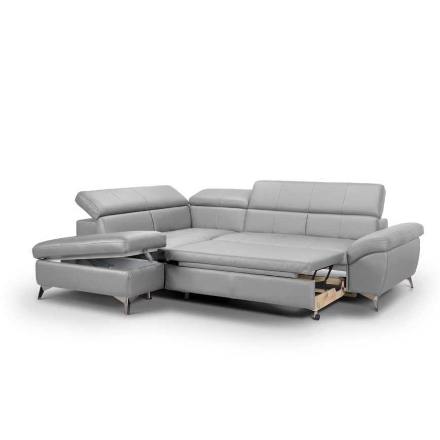 Rohová sedačka svetlosivej kože je rozložená, odhalený je úložný priestor. Súčasťou je aj samostatný taburet s úložným priestorom. Sedačka stojí na svetlom pozadí.