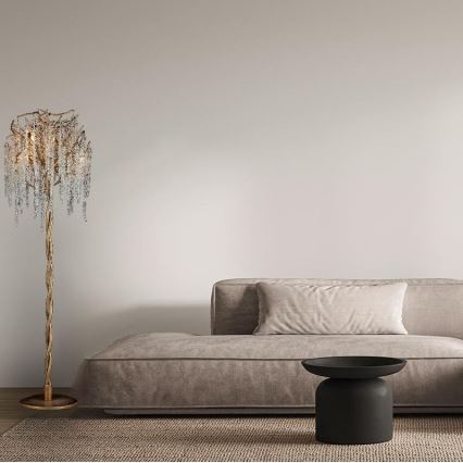 Stojacia lampa v tvare stromu s krištáľovými príveskami svieti v obývacej izbe. Vedľa nej sa nachádza hnedá pohovka s vankúšom a nízky čierny stolík.