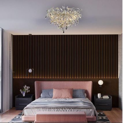 Luster svieti v spálni. Je inštalovaný na strope nad ružovou posteľou s posteľnou bielizňou a vankúšmi. Stena za posteľou je tmavohnedá s vertikálnymi lamelami.