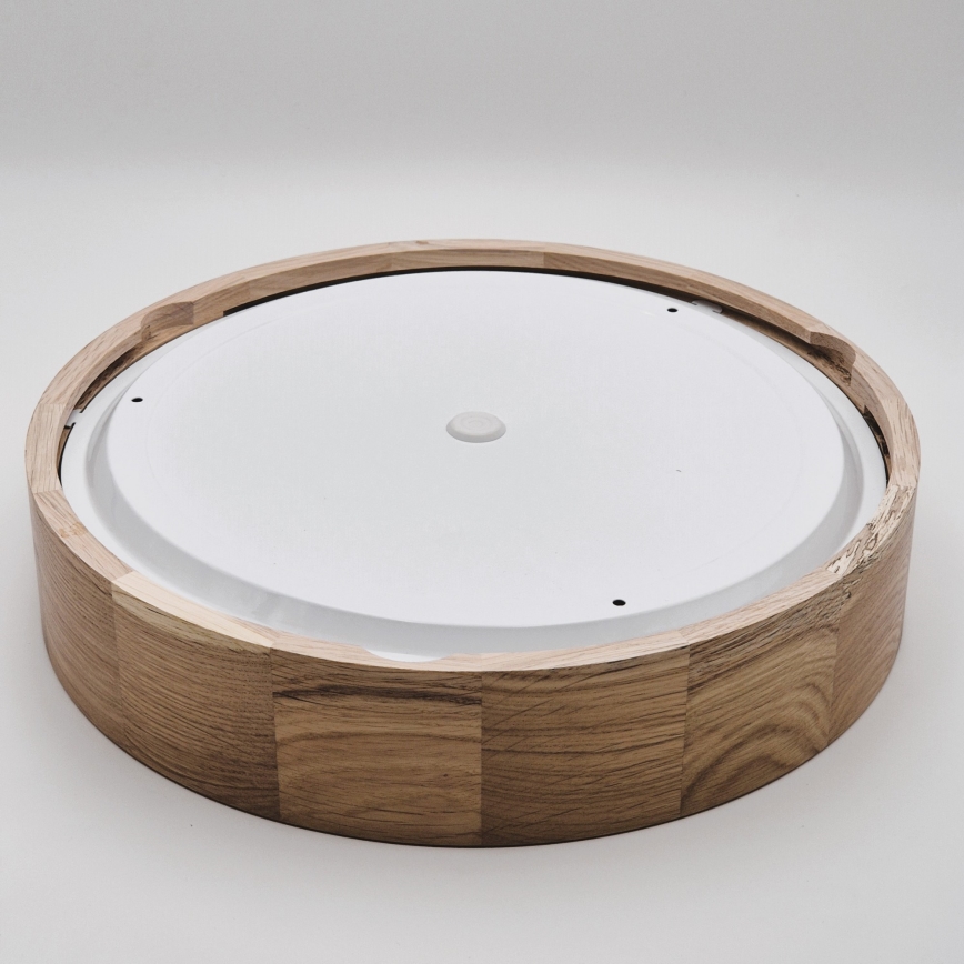 Drevené svietidlo s bielym difúzorom je položené na svetlom podklade. Svietidlo je zložené z kruhového dreveného rámu a vnútorného bieleho kruhu. Rám je z viditeľných kusov dreva.