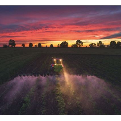Traktor postrekuje pole pri západe slnka. Svetlo z reflektorov osvetľuje rozprašovač a rastliny. Na obzore sa rozprestiera dramatická, červeno-oranžová obloha.