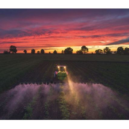 Traktor postrekuje pole pri západe slnka. Na obzore sú viditeľné stromy. Svetlo slnka maľuje oblohu odtieňmi červenej a oranžovej.