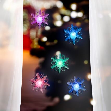 Štyri snehové vločky s LED svetlami sú pripevnené na okne. Vločky vyžarujú farebné svetlo. Za oknom je rozostrené vianočné osvetlenie.