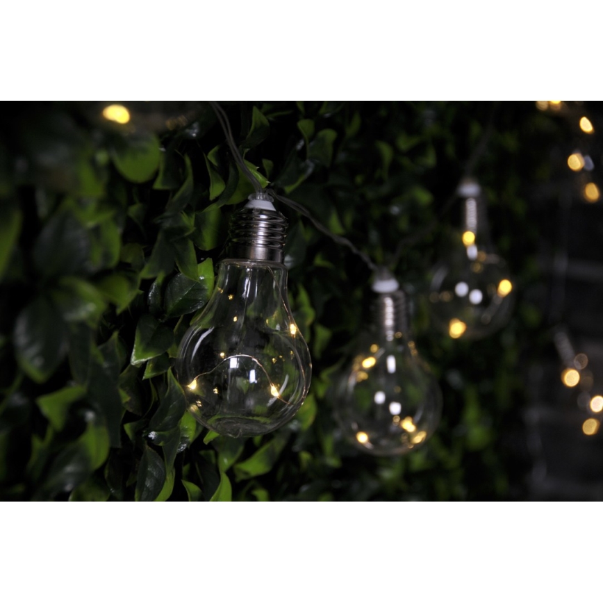 Sklenené žiarovky svietia na svetelnom reťazi. Žiarovky sú zavesené na tmavozelenej umelej stene. Svetlo vyžaruje teplú žltú farbu.