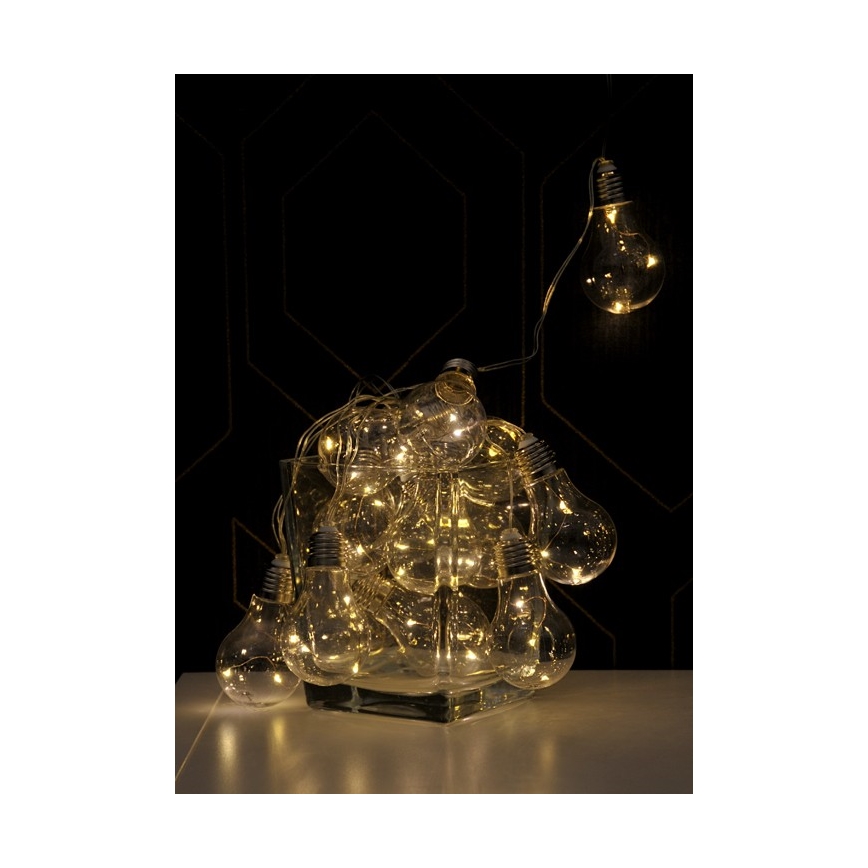 Svetelný reťaz s LED žiarovkami v tvare klasických žiaroviek je umiestnený v sklenenej váze. Žiarovky svietia teplým svetlom. V pozadí je tmavá stena s geometrickým vzorom.