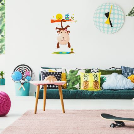 Opica visí dolu hlavou z konára stromu na nástenných dekoráciách v detskej izbe. Na gauči sú vankúše s tropickými motívmi. V miestnosti sa nachádzajú hračky a nábytok pre deti.