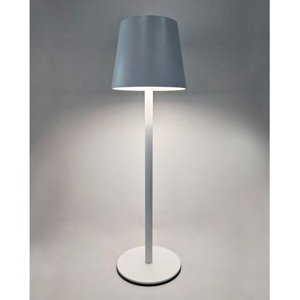 Stojacia lampa svieti. Má šedý tienidlo v tvare zrezaného kužeľa a bielu kovovu nohu s okrúhlou základňou. Je umiestnená na svetlom pozadí.