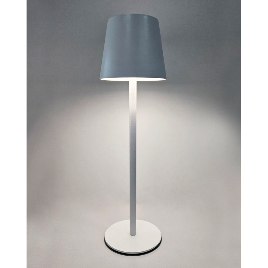 Stojacia lampa svieti. Má šedý tienidlo v tvare zrezaného kužeľa a bielu kovovu nohu s okrúhlou základňou. Je umiestnená na svetlom pozadí.