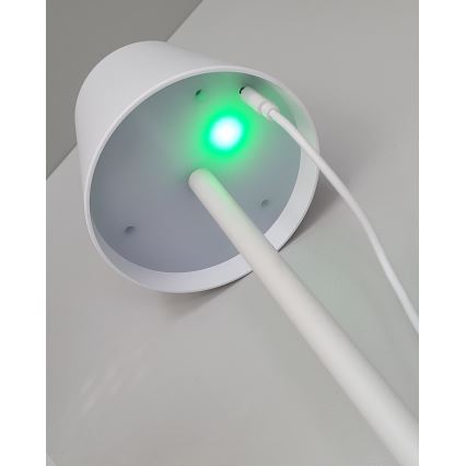 Biela stojaca lampa s valcovou základňou a tenkým stĺpikom má v spodnej časti zelené svetlo. K základni sú pripojené dva biele káble. Lampa stojí na svetlom povrchu.
