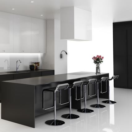 Kreslo stojí pri stene s geometrickým vzorom. Svietidlo na stene osvetľuje kreslo. V pozadí je drevená podlaha a nábytok.