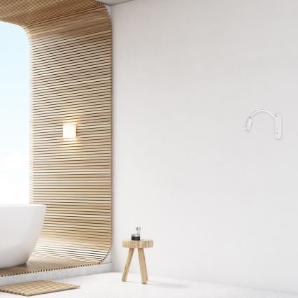 Biela nástenná lampa s ohybným ramenom je umiestnená na bielej stene. Nachádza sa v kúpeľni s drevenou stenou a voľne stojacou vaňou. V blízkosti je malý drevený stolík.