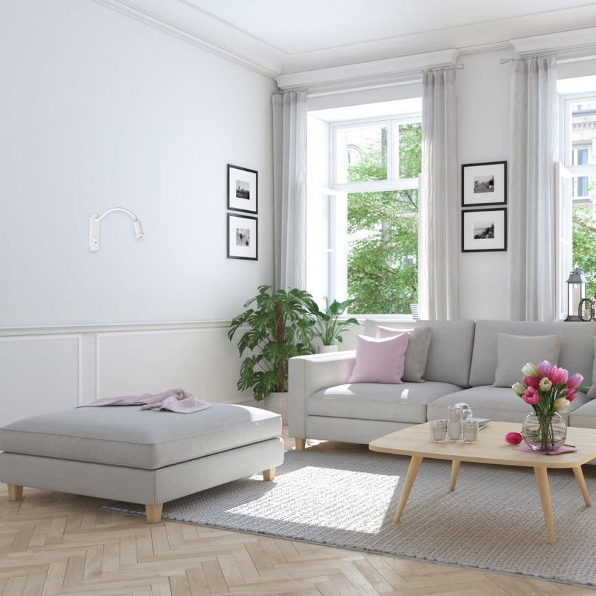 Nástená lampa svieti v obývacej izbe. Svetlo dopadá na svetlosivú sedačku a konferenčný stolík s kvetmi. V miestnosti je svetlosivá pohovka, koberec a drevená podlaha.