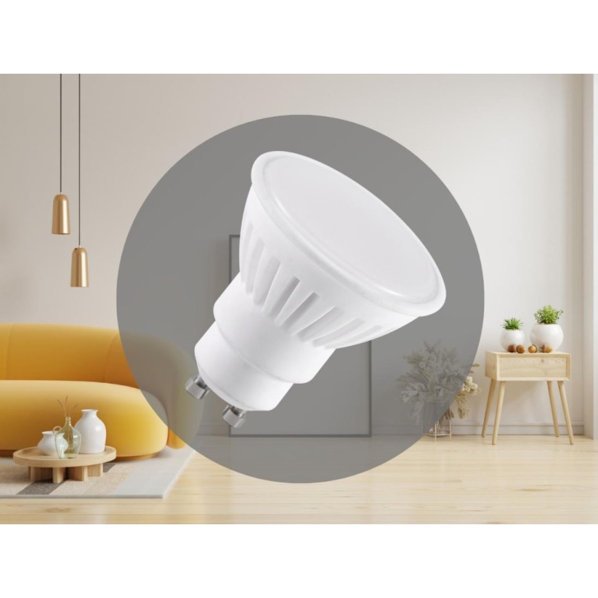 LED žiarovka GU10 je zobrazená v interiéri. Žiarovka je biela, s plochým vrchom a držiakom GU10. Nachádza sa v obývacej izbe s nábytkom a doplnkami.