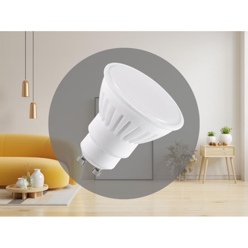 LED žiarovka je zobrazená v interiéri obývacej izby. Je biela, guľato-kužeľovitého tvaru so závitom. Žiarovka je umiestnená v sivej kruhovej ploche na pozadí nábytku, podlahy a stien.