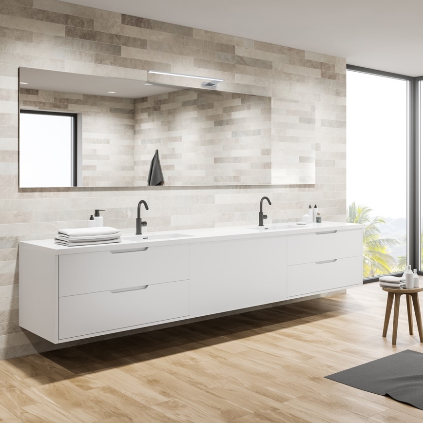 Dvojité umývadlo s dvoma batériami je umiestnené na bielej nástenní skrinke. Skrinka je zavesená na stene obloženej svetlohnedými dlaždicami. Nad umývadlom je veľké zrkadlo s integrovaným osvetlením.